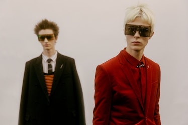 A model in a red suit and square sunglasses standing in front of a model in a black blazer and strip...