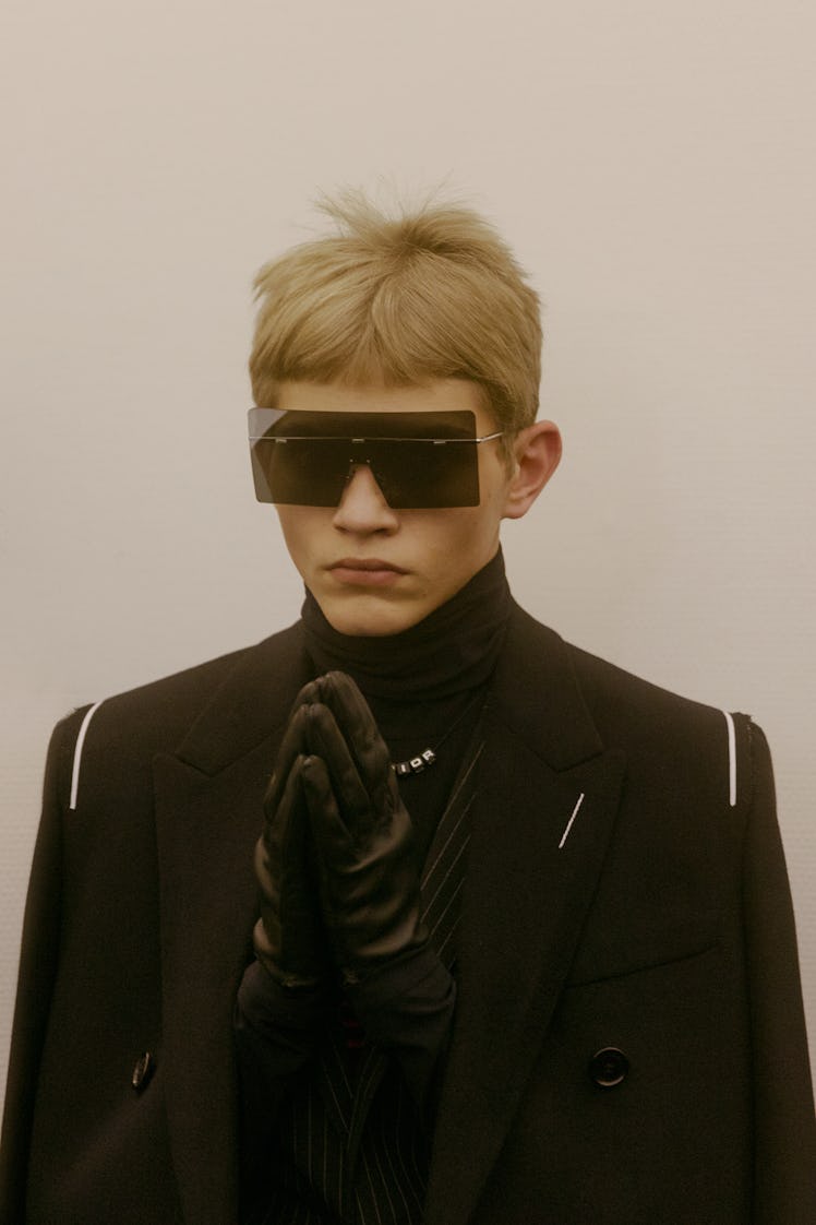 A model in a black coat, turtleneck, gloves and sunglasses holding his hands together backstage at t...