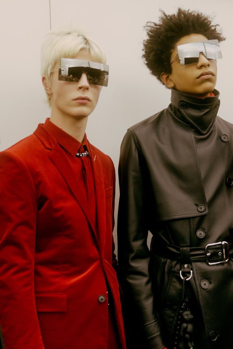 A model in a red suit standing next to a model in long black leather trench coat at the Dior Homme F...