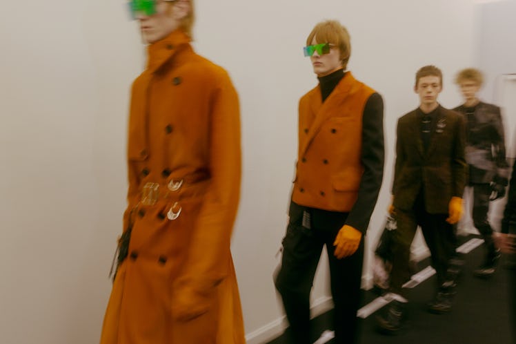A model in an orange trench coat, followed by other models, walking towards the runway at the Dior s...