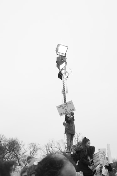 A person climbing onto a tall stand with two reflectors on top and holding a protest sign, the Women...