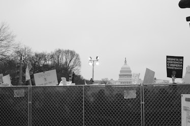 A group of people with protest posters semi-visible behind a fence and the US Capitol in the backgro...