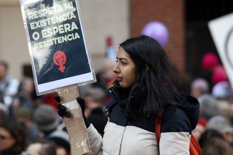 A woman in a black-white jacket and holding a protest poster 'Respeta mi existencia o espera resiste...