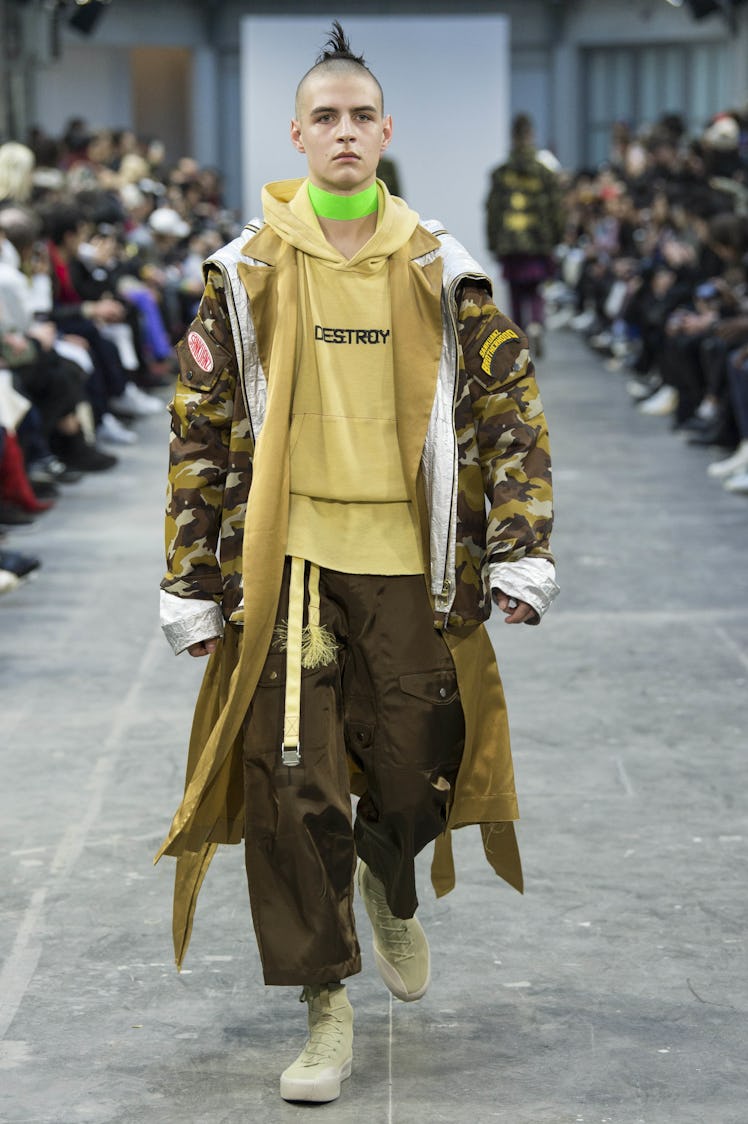 A model wearing brown pants, a beige hoodie, and a camo jacket with a green choker at the Sankuanz F...