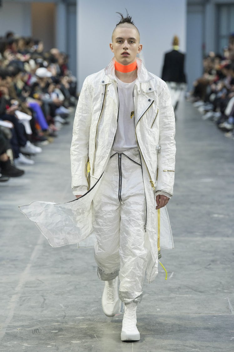 A model wearing a white jumpsuit, and a long white jacket with an orange choker at the Sankuanz Fall...