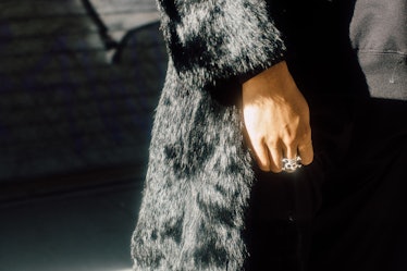 A close-up of a person in a black fur coat, wearing a rhinestone skull ring on their middle finger a...