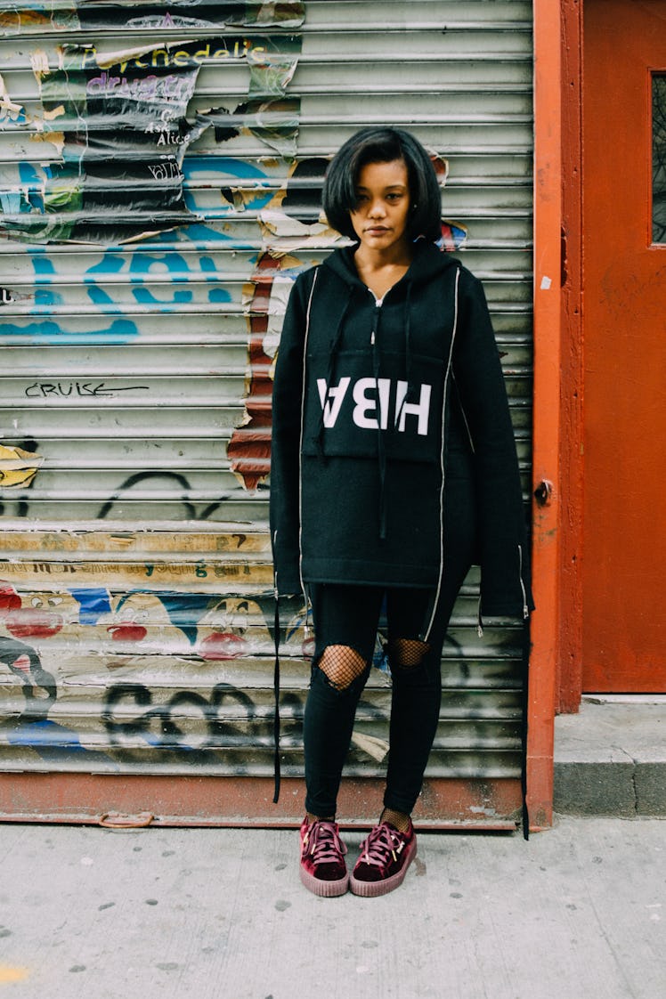 A woman in a black hoodie with the upside-down text ABH, black trousers and burgundy sneakers