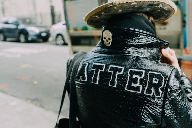 The back of a woman in a black leather jacket with the text ATTER and a beige hat at Hood by Air