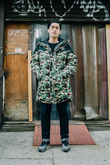 A man standing in a camo print puffer jacket, black pants and grey sneakers