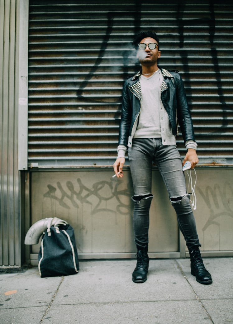 A man standing in a beige shirt, black leather jacket, grey denim jeans and black boots 