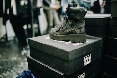 A khaki-brown boot placed on a black box and people blurred in the background at Hood by Air