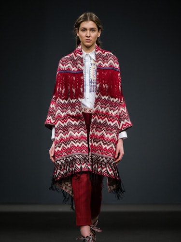 A model wearing a red-white patterned fringed coat and red trousers on the runway