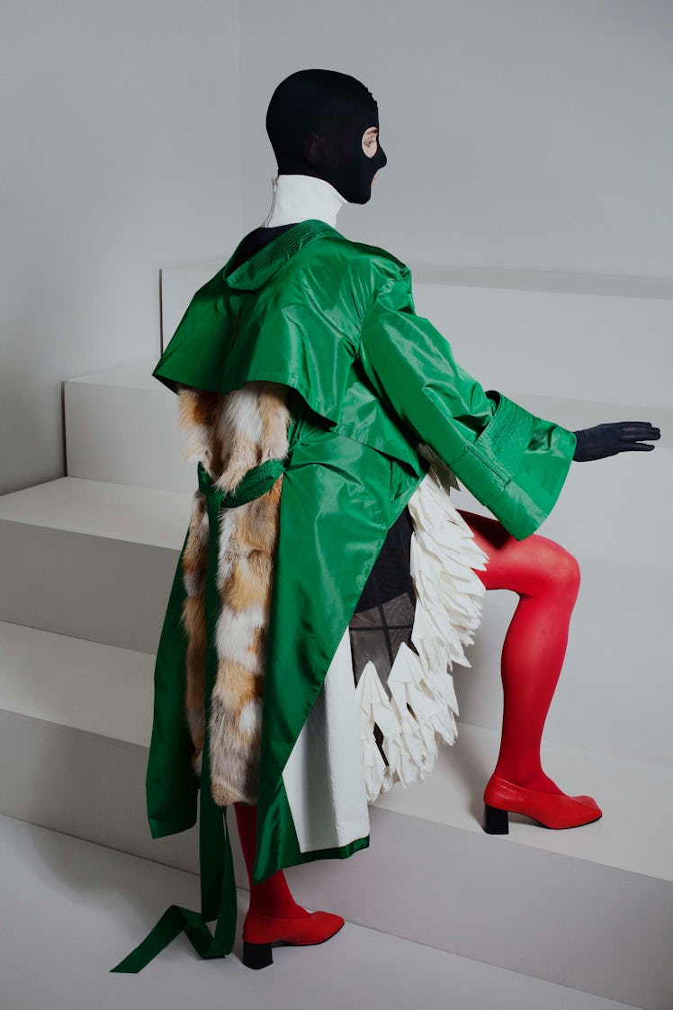 A model in a green trench coat, red tights, red shoes and a black ski mask from the unisex collectio...