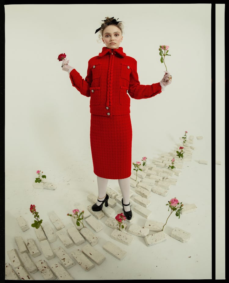 A woman in a red skirt-suit holding two roses while the rest is placed on the brick path on which sh...