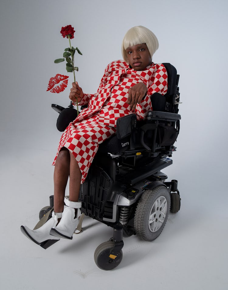 A woman in a wheelchair, wearing a red-and-white checked skirt suit paired with white boots, holding...