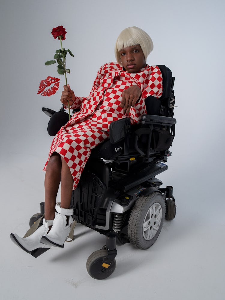 A woman in a wheelchair, wearing a red-and-white checked skirt suit paired with white boots, holding...