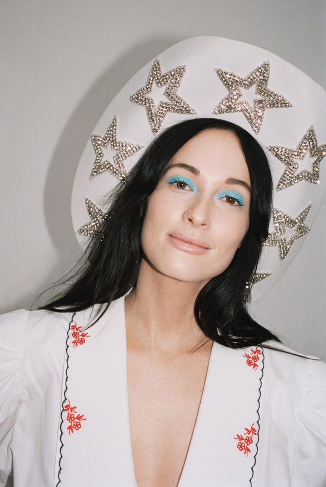 A brunette with blue eyeshadow wearing a white hat adorned with rhinestone stars and a white shirt