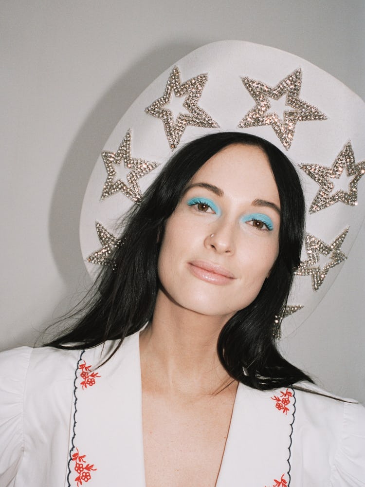 A brunette with blue eyeshadow wearing a white hat adorned with rhinestone stars and a white shirt
