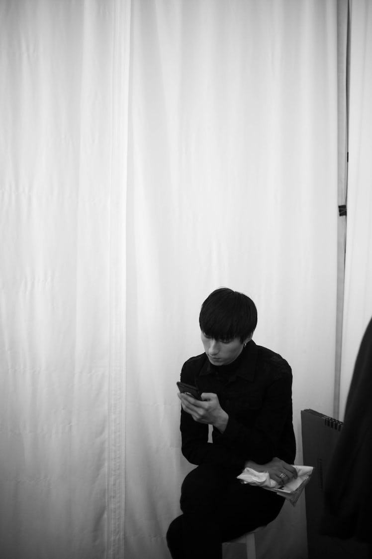 A model in a black turtleneck and black pants scrolling on his phone backstage at the Zadig & Voltai...