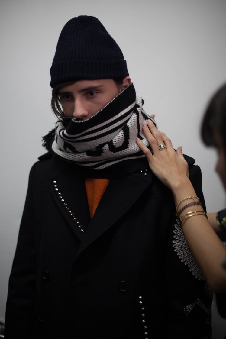 A model in a black beanie getting an oversized black and white scarf put on him backstage at the Zad...