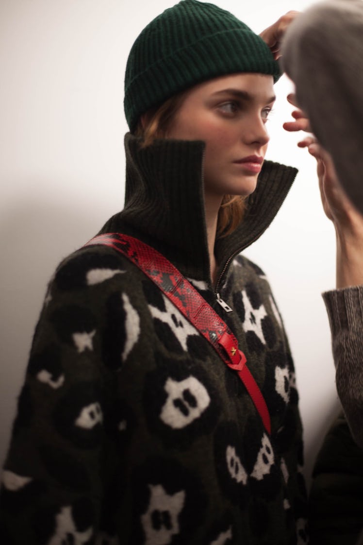 A model getting dressed in a green beanie and a black fleece jacket with white illustrations on it 