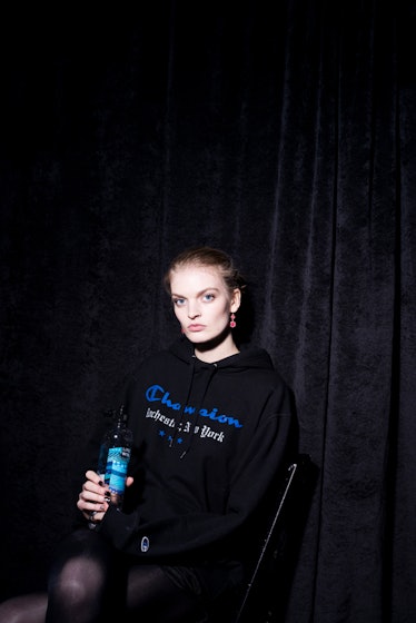 A model sitting in a black hoodie with print while holding a drink backstage at Monse Fall 2017