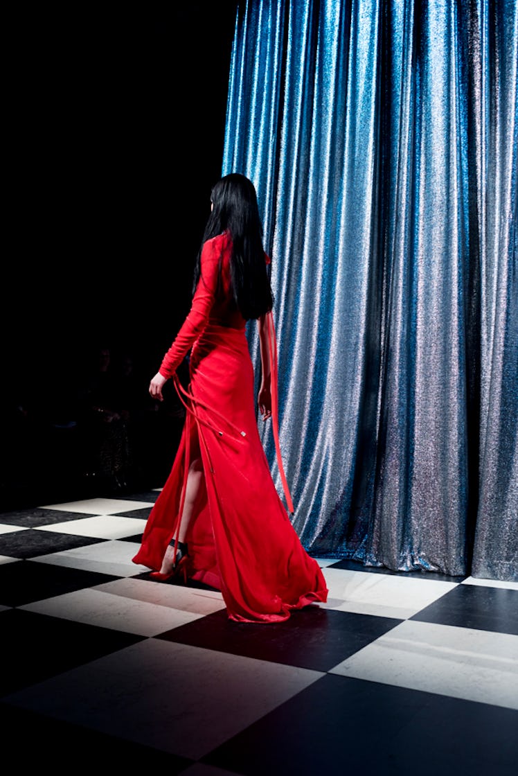 A model with long black hair walking in a red dress backstage at Monse Fall 2017