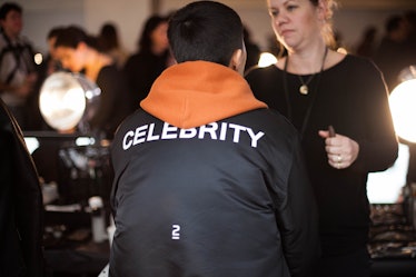 A male model sitting while wearing a black jacket with a "celebrity" text sign on the back