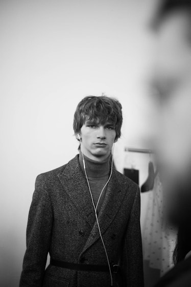 A male model walking while wearing a grey coat and turtleneck sweater 