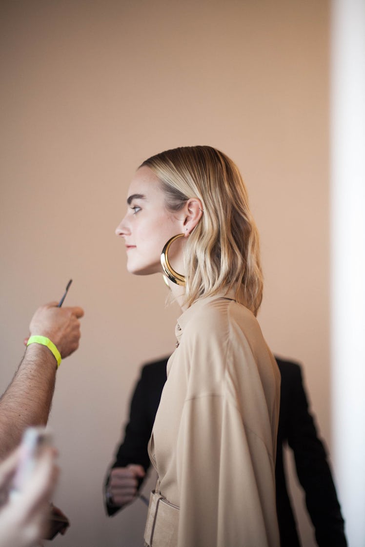 A makeup artist putting makeup on a face of a blonde woman with round golden earrings