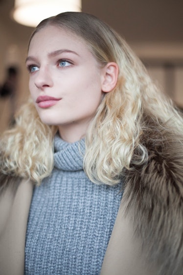 A blonde model posing in a grey turtleneck sweater and in a brown coat
