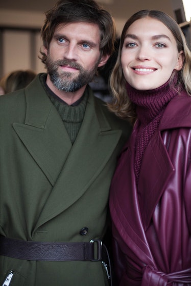 A man posing in a green coat and sweater combination next to a woman with a purple coat and sweater ...