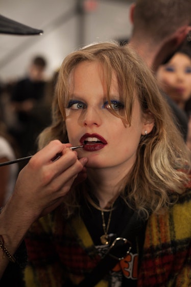 A makeup artist applying burgundy lipstick on a blonde model backstage at Anna Sui Fall 2017