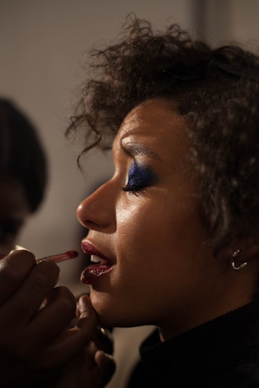 A makeup artist applying burgundy lipstick on a model with blue eyeshadow backstage at Anna Sui Fall...