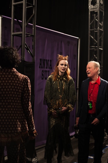 A model in a green dress and a double top knot standing and talking to a man backstage at Anna Sui F...