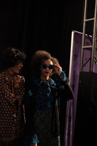 A model in a brown geometric patterned suit and a model in a black-blue coat and dress  backstage at...