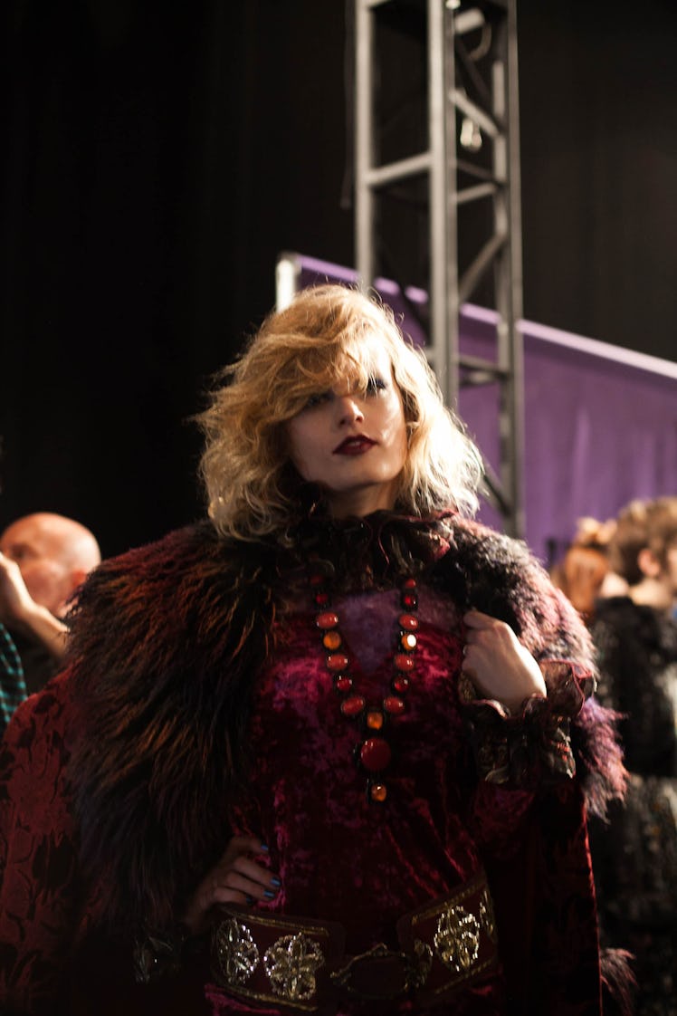 A blonde model in a pink dress, orange beaded necklace and a burgundy fur coat backstage at Anna Sui...