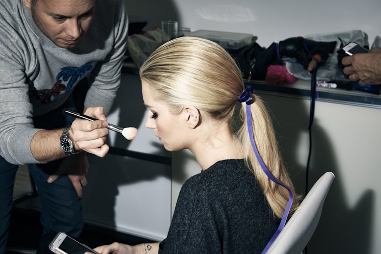 Lottie Moss typing on her phone while a makeup artist is putting makeup on her face