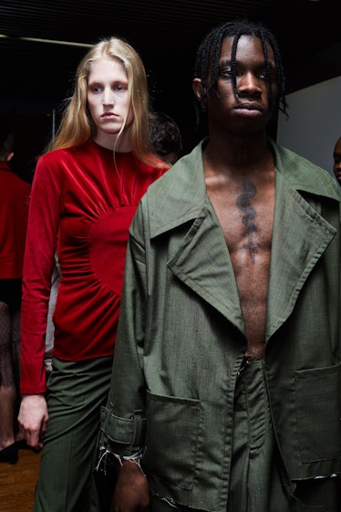 A male model standing while wearing a green coat and a blonde female model standing while wearing a ...