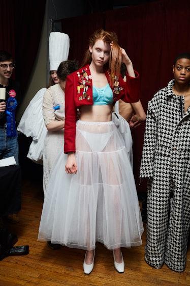 A female model wearing a red blazer, light blue bra, and a white dress over white underpants