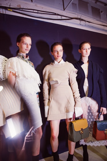Three female models standing while wearing white mini dresses and holding handbags at the backstage ...