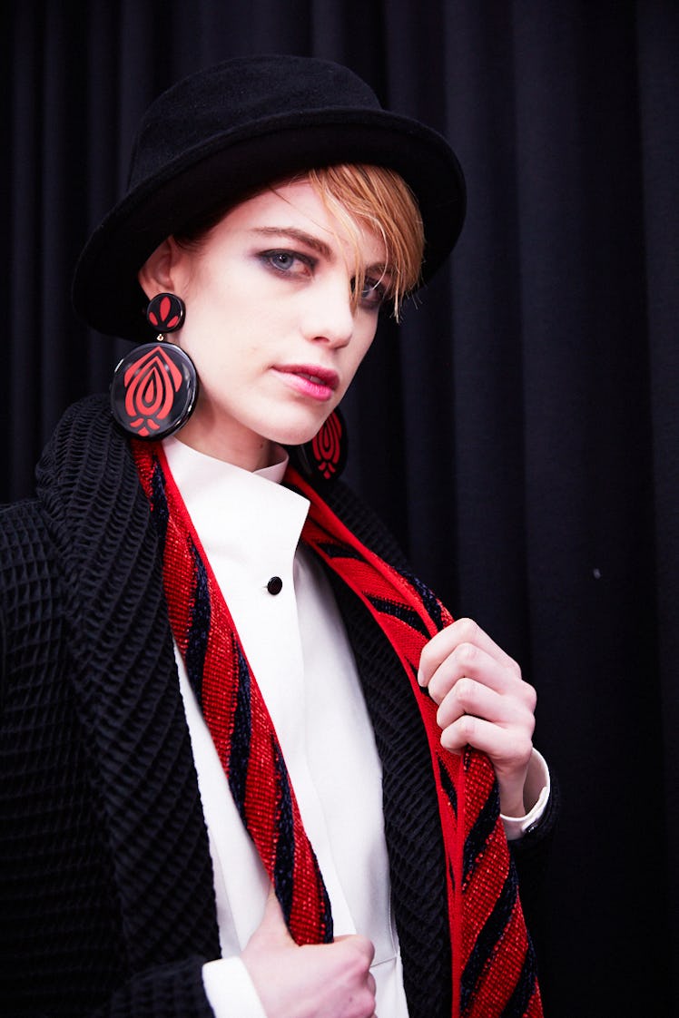 A blonde lady posing in a black blazer, black hat, and white shirt while holding a red and black tie...