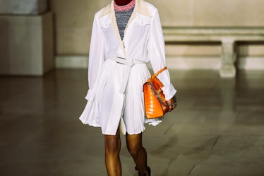 A model in a white shirtdress, holding an orange bag walking the runway for Louis Vuitton’s Fall 201...