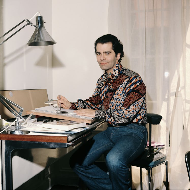 Karl Lagerfeld sitting at a design desk with a pen in a lush silk shirt and denim jeans.