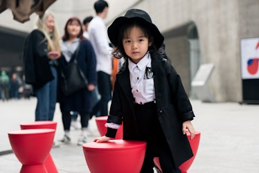 A toddler wearing a black hat, pants and a trench coat over a white shirt on the streets of Seoul.