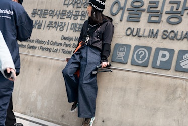A woman wearing wide pants, a black jacket and a beanie while leaning on the wall in Seoul