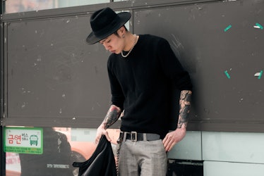 A man wearing a black t-shirt, grey pants and a black hat standing next to a wall in Seoul.