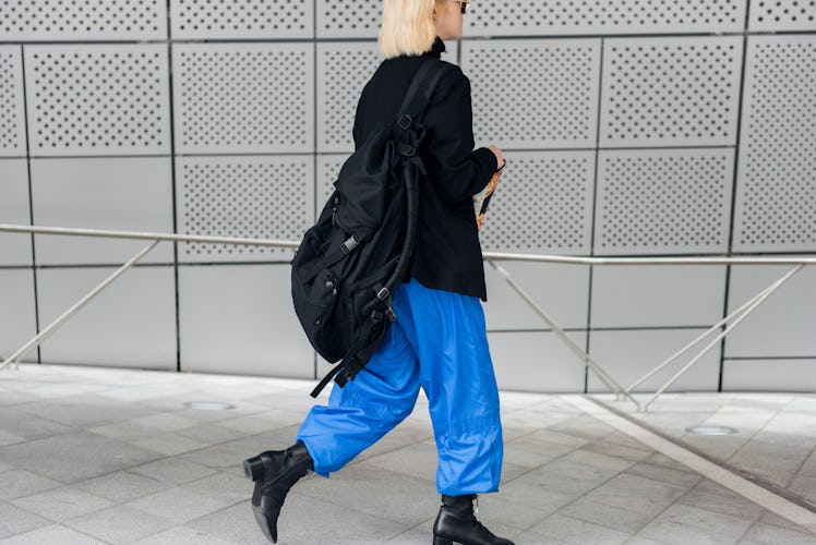 A woman wearing a black blazer, wide blue pants and a black backpack while walking.