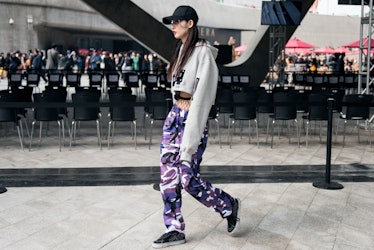 A girl wearing purple camouflage pants with fishnets under, a white cropped hoodie, and a black cap ...