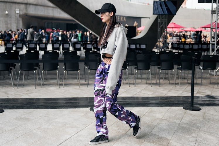 A girl wearing purple camouflage pants with fishnets under, a white cropped hoodie, and a black cap ...